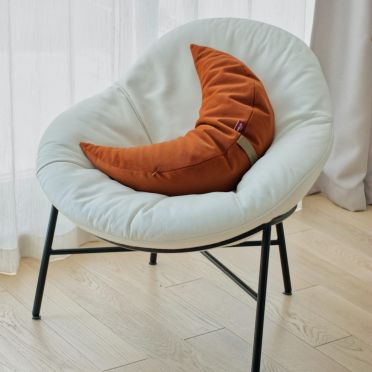 poduszka w kształcie księżyca w kolorze rudym, ciemno pomarańczowym leży na białym, dużym fotelu typu papassan. drewniana podłoga, spokojny, jasny wystrój.